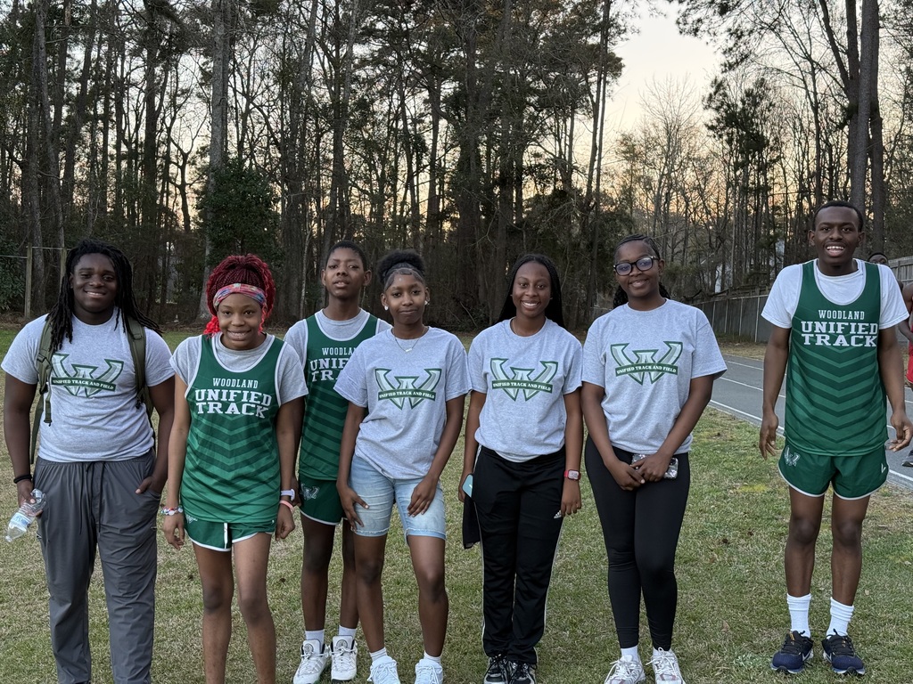 A group of Woodland Unified Track athletes walk toward the camera along a grassy area beside a track, smiling and relaxed. They wear a mix of green uniforms and gray team shirts, with trees and a road in the background during early evening light.