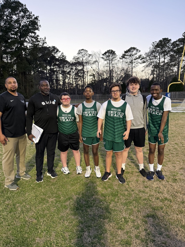 Woodland Unified Track athletes stand in a line on a grassy field with two coaches beside them. The students wear green uniforms, and the coaches are dressed in black athletic gear. A track, goalpost, and tree line are visible behind them under the evening sky.