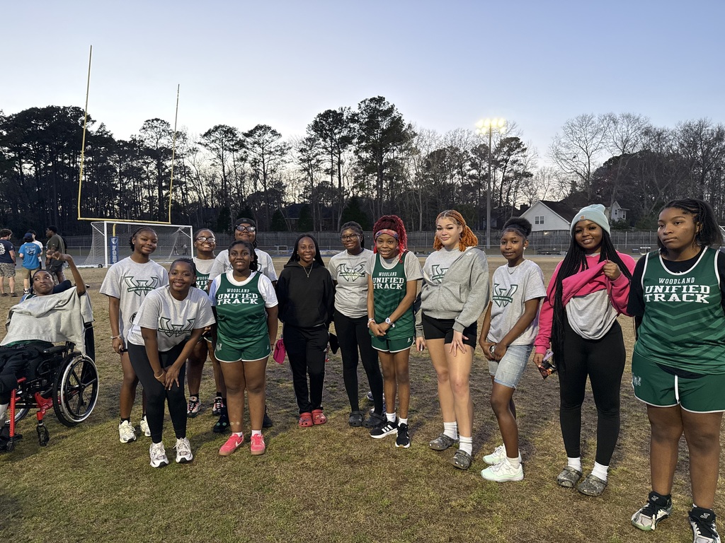 A large group of Woodland Unified Track athletes pose together on a grassy field near a track and football goalpost at dusk. Students wear green and gray “Woodland Unified Track” uniforms and T-shirts, smiling and standing shoulder to shoulder. A student in a wheelchair is visible on the far left, and stadium lights glow in the background with trees lining the field.