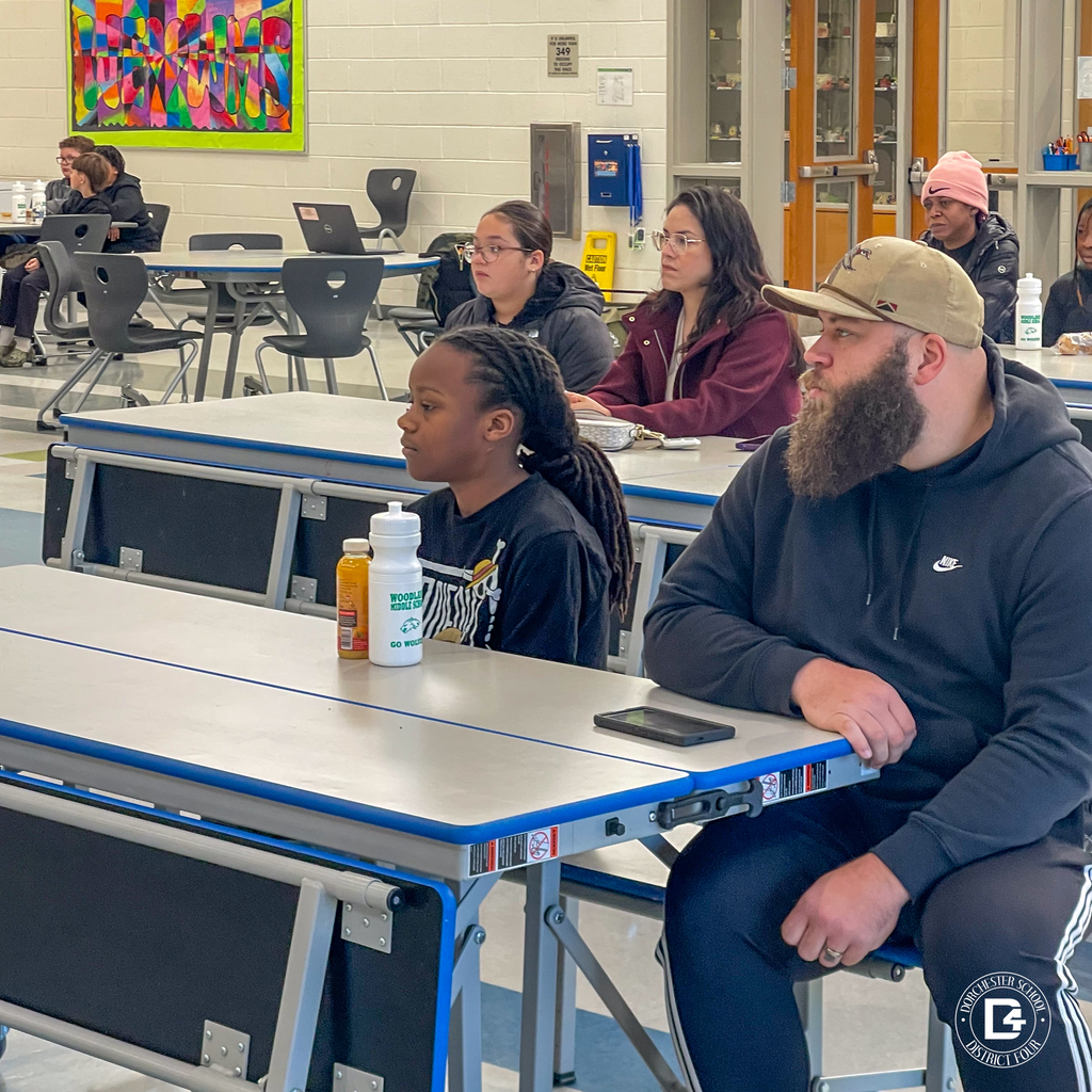 Parents and students sit together at cafeteria tables, listening attentively during an informational session about transitioning to Woodland Middle School.