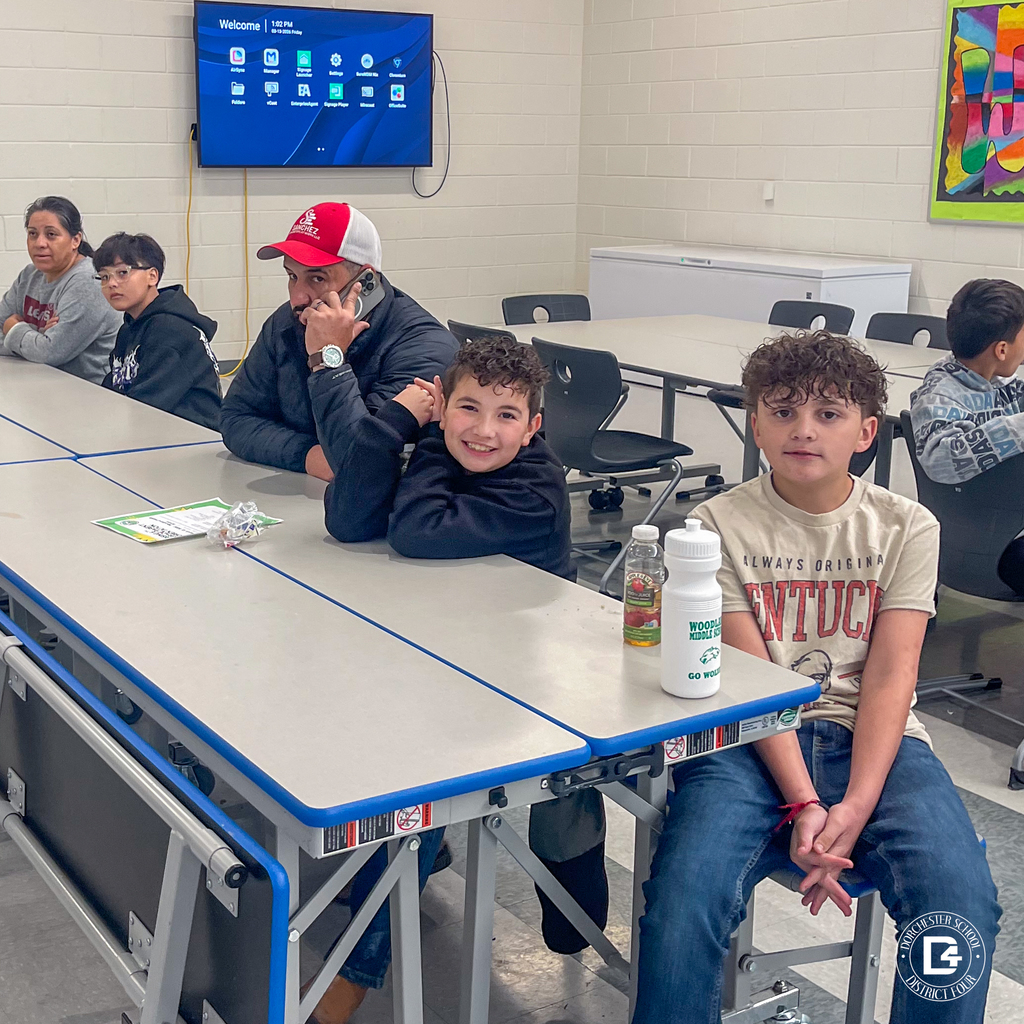 Parents and students sit at tables in the cafeteria while listening to a presentation, with informational materials and drinks placed in front of them.