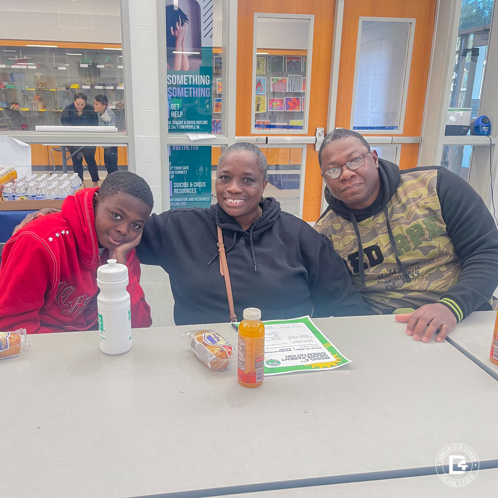 A family of three sits together at a cafeteria table, smiling for the camera with refreshments and event information in front of them.