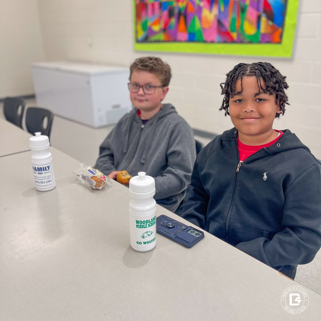 Two students sit at a cafeteria table with snacks and water bottles, smiling as they participate in the Rising 6th Grade Parent Open House.
