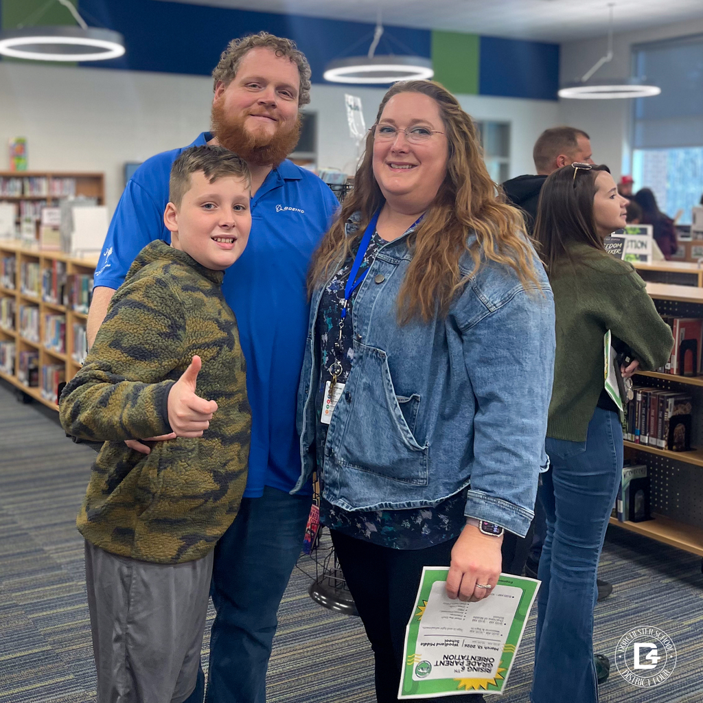 A family poses together inside the school library, smiling during the Rising 6th Grade Parent Open House as they prepare for the upcoming school year.