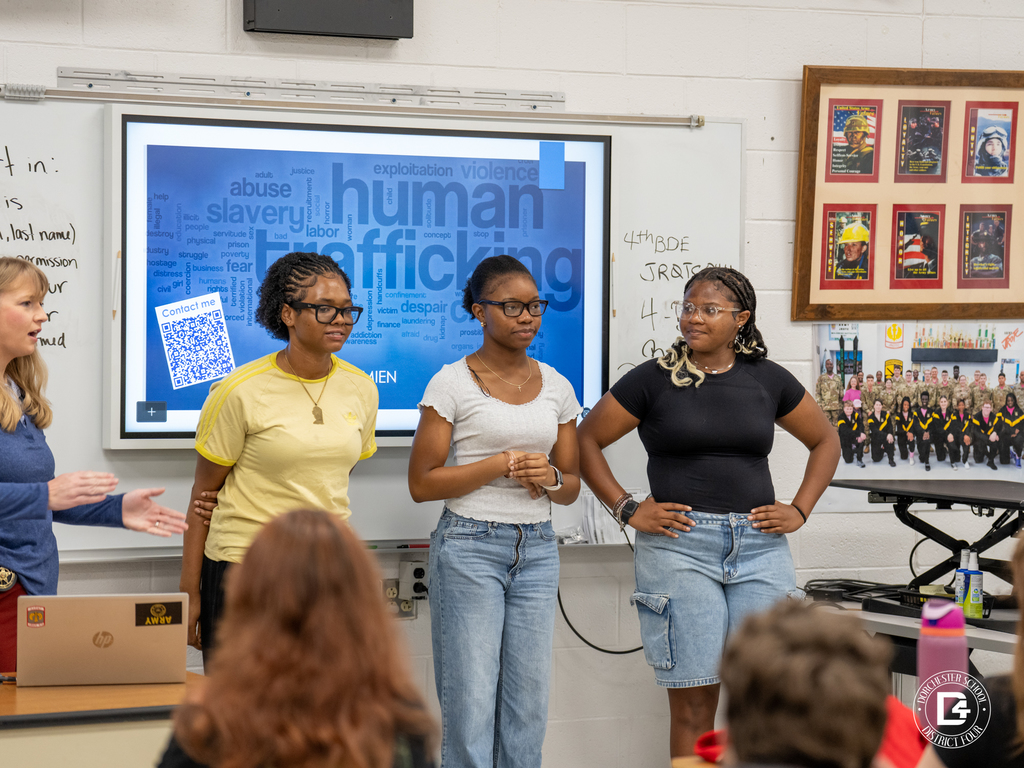 Three FCCLA students stand at the front of the classroom during a presentation while the guest speaker gestures beside them, with a human trafficking awareness slide visible behind them.