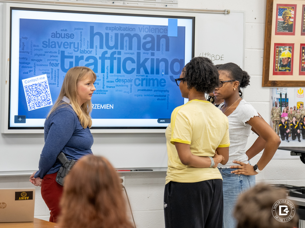 A law enforcement officer speaks with two students in a classroom at Woodland High School, standing in front of a screen displaying information about human trafficking awareness.