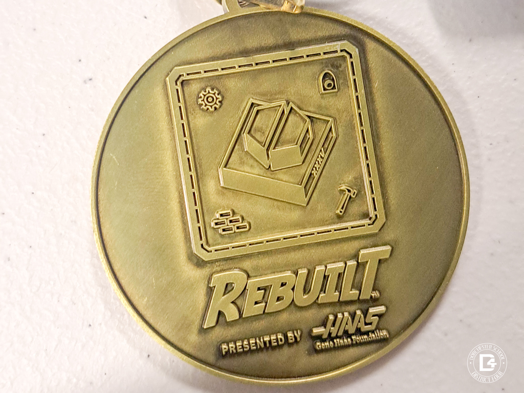 Close-up of a gold FIRST Robotics Competition medal labeled “Rebuilt” presented by Haas, featuring icons of tools and materials.