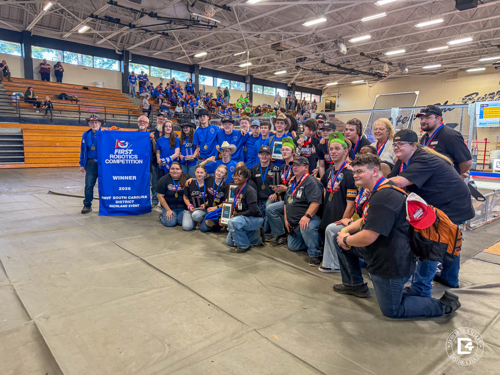 Group of students and mentors pose on a gym floor at a robotics competition, many wearing medals and team shirts, smiling and holding a horse prop in front of their robot setup.