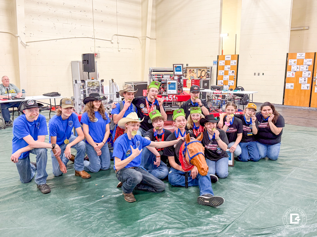 Group of students and mentors pose on a gym floor at a robotics competition, many wearing medals and team shirts, smiling and holding a horse prop in front of their robot setup.
