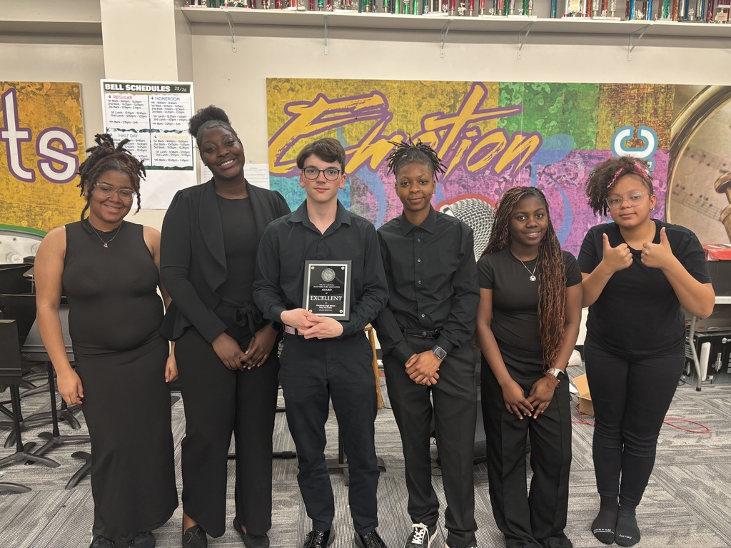 Six Woodland High School students dressed in black concert attire stand together in a music classroom, with one student in the center holding an “Excellent” award plaque. Colorful wall art and a row of trophies are visible behind them, highlighting the band program’s achievements.