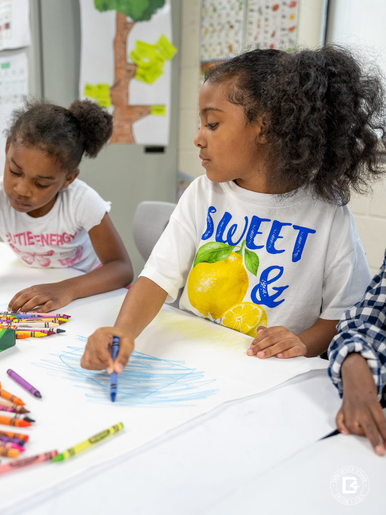 Student carefully drawing on blue paper with crayons while classmates work nearby.