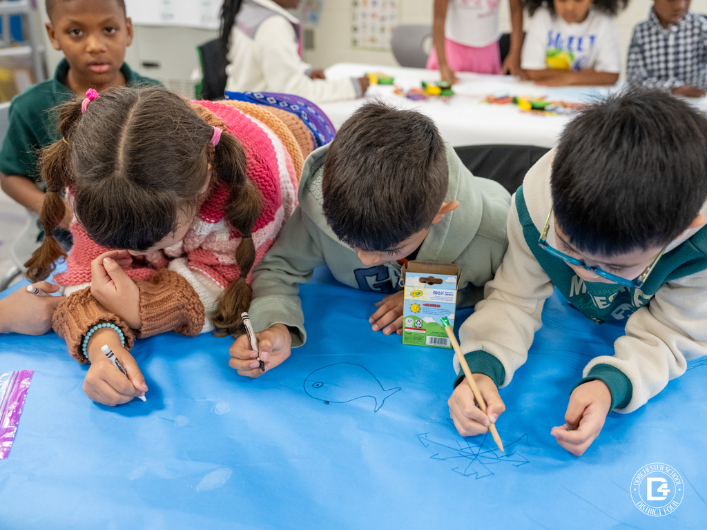 Students leaning over a table working together to draw ocean scenes on large blue paper with crayons.