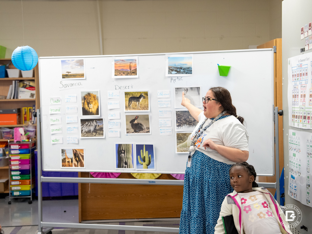 Teacher standing beside a classroom board pointing to images of ecosystems labeled savanna, desert, and Arctic while students observe.