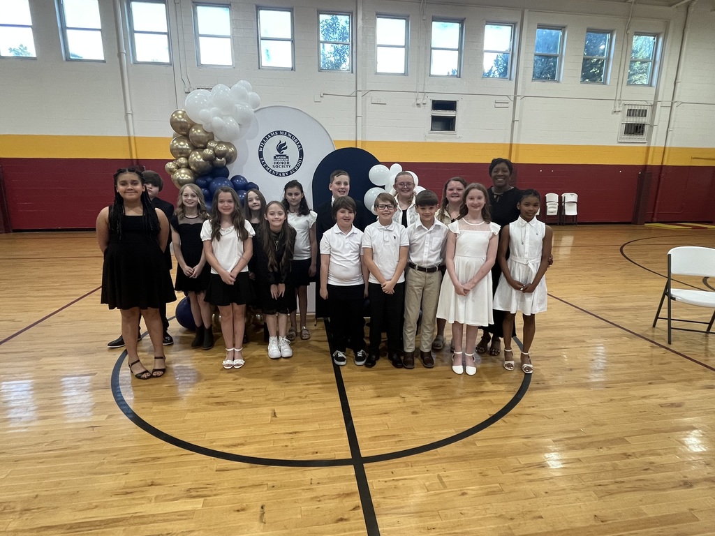 A group of elementary students dressed in black and white attire stand in a school gymnasium for a National Elementary Honor Society induction ceremony. They are posed in front of a decorative balloon display in white, gold, and navy, with a circular NEHS backdrop. A few adults stand behind the students, and the gym’s red and gold walls and high windows are visible in the background.