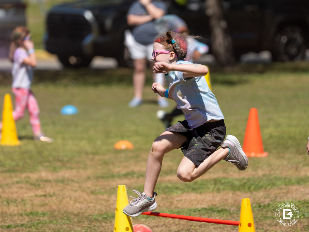 Student mid-jump over a hurdle during the Clay Hill Elementary Color Fun Run, running through a cone-marked course on the field.