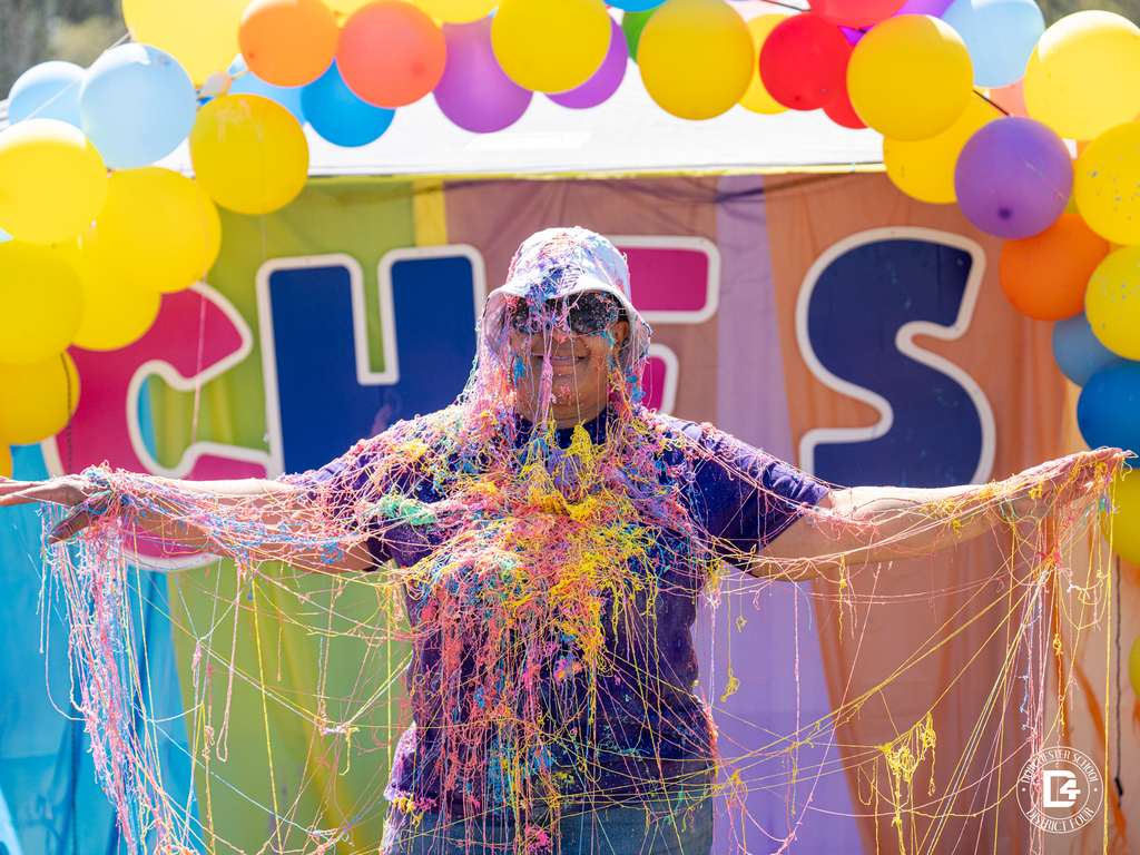 Adult standing under a colorful balloon arch at the Clay Hill Elementary Color Fun Run, covered head to toe in bright string spray with arms outstretched in celebration.