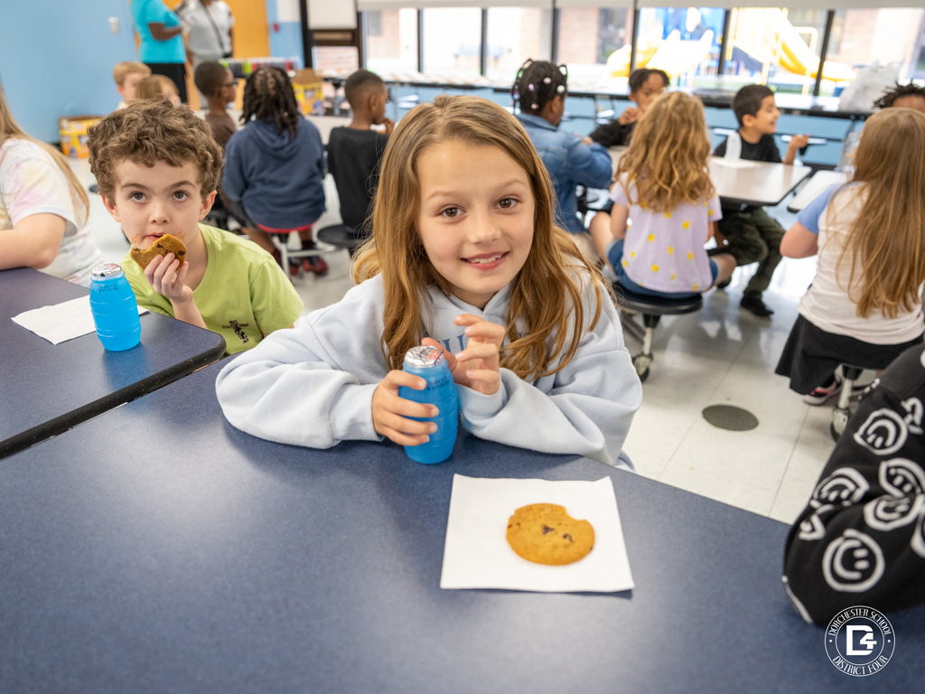 A young elementary student smiles at the camera while holding a blue juice drink next to a chocolate chip cookie on a napkin, celebrating her Quarter 3 attendance achievement in the school cafeteria.