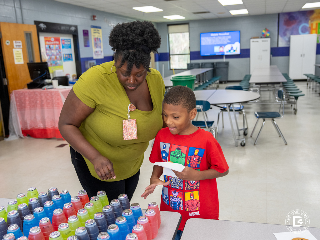 A school staff member wearing a lanyard helps a young student in a red Marvel t-shirt select a juice drink from a table filled with colorful juice bottles during a student attendance recognition event.
