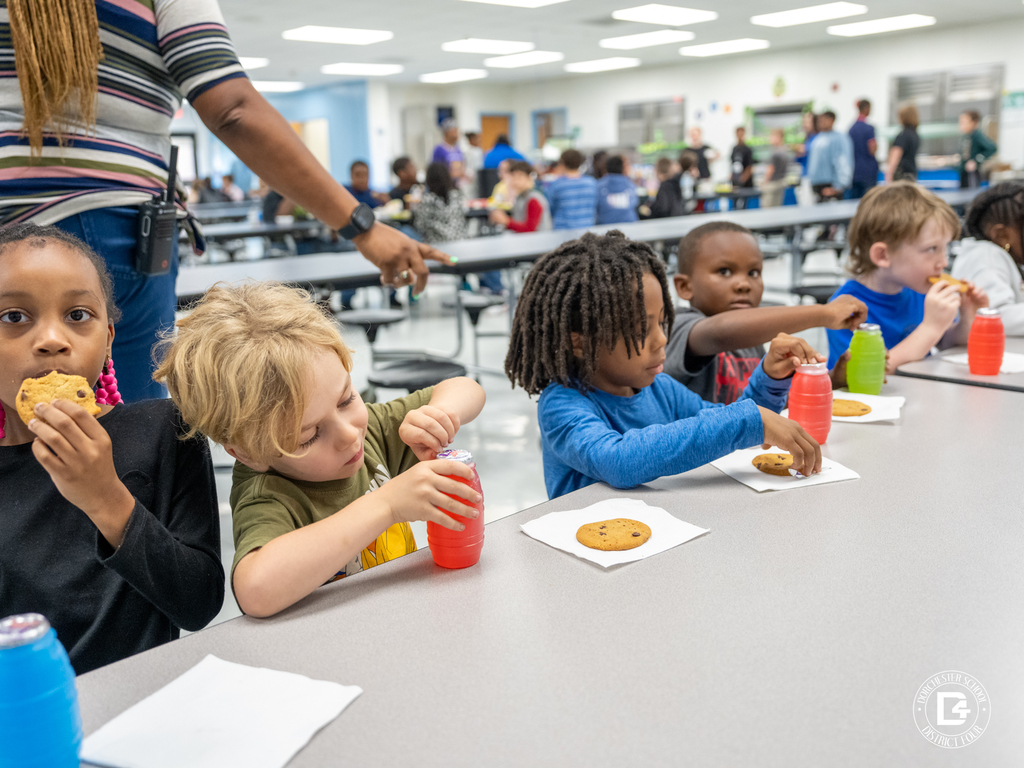 A group of young elementary students sit together at a cafeteria table enjoying chocolate chip cookies and colorful juice drinks during a Quarter 3 attendance celebration at Dorchester School District Four, with a packed cafeteria of students visible in the background.