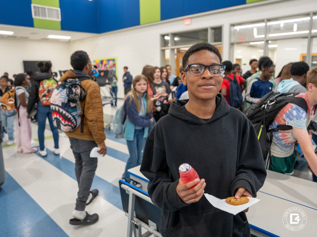 A middle school student wearing glasses and a black hoodie holds a red juice drink and a cookie on a napkin while students line up behind him during a Quarter 3 attendance recognition event at Dorchester School District Four.