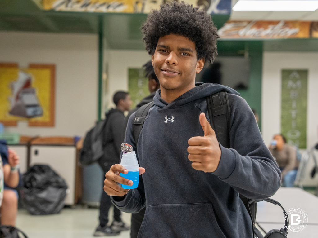 A high school student in a black Under Armour hoodie smiles and gives a thumbs up while holding a blue juice drink, celebrating his Quarter 3 attendance achievement at Dorchester School District Four.