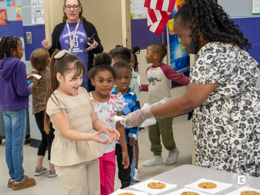 A school staff member wearing gloves hands out cookies to a line of excited elementary students in a school cafeteria while a teacher supervises in the background during a Quarter 3 attendance celebration.