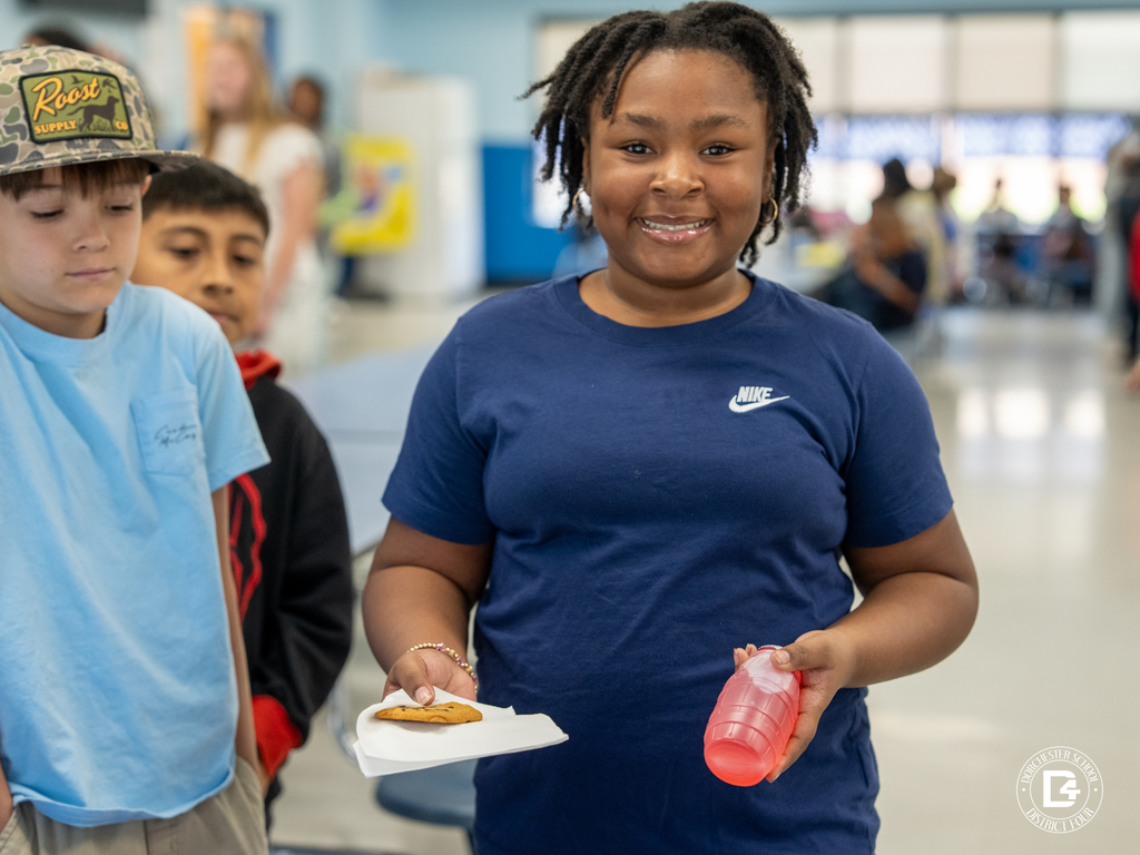 A smiling elementary student in a navy Nike t-shirt holds a chocolate chip cookie on a napkin and a red juice drink while walking through the school cafeteria during a Quarter 3 attendance celebration at Dorchester School District Four.