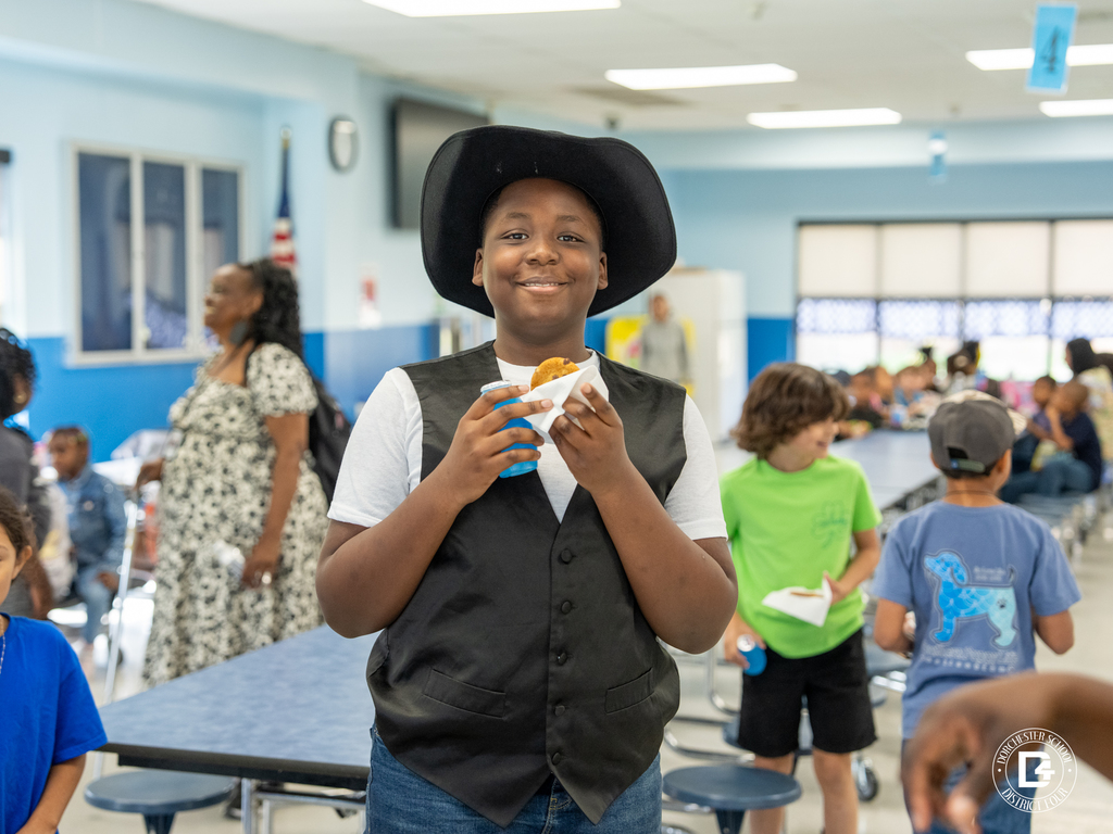 A smiling elementary school student wearing a black cowboy hat and vest holds a snack in a school cafeteria during a Quarter 3 attendance celebration at Dorchester School District Four.