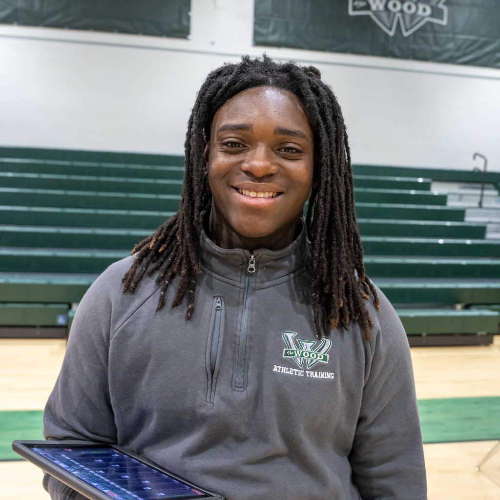 Student standing in a gymnasium in front of green bleachers, smiling and holding a tablet. He is wearing a gray Woodland Athletic Training pullover with the school logo visible on the chest.