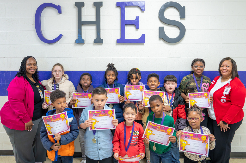A group of Clay Hill Elementary School students stand in front of a wall with large “CHES” letters, each holding a “Student of the Month” certificate. The students, wearing medals, are joined by two staff members on either side, smiling and celebrating their achievement. The group represents a diverse set of young learners recognized for their hard work and positive behavior.
