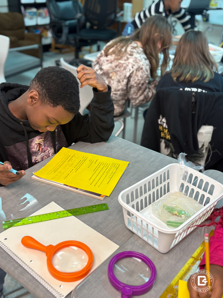 A student focuses on a yellow worksheet at a classroom table with magnifying tools and a basket holding a frozen sample. Other students sit nearby collaborating on the same activity.