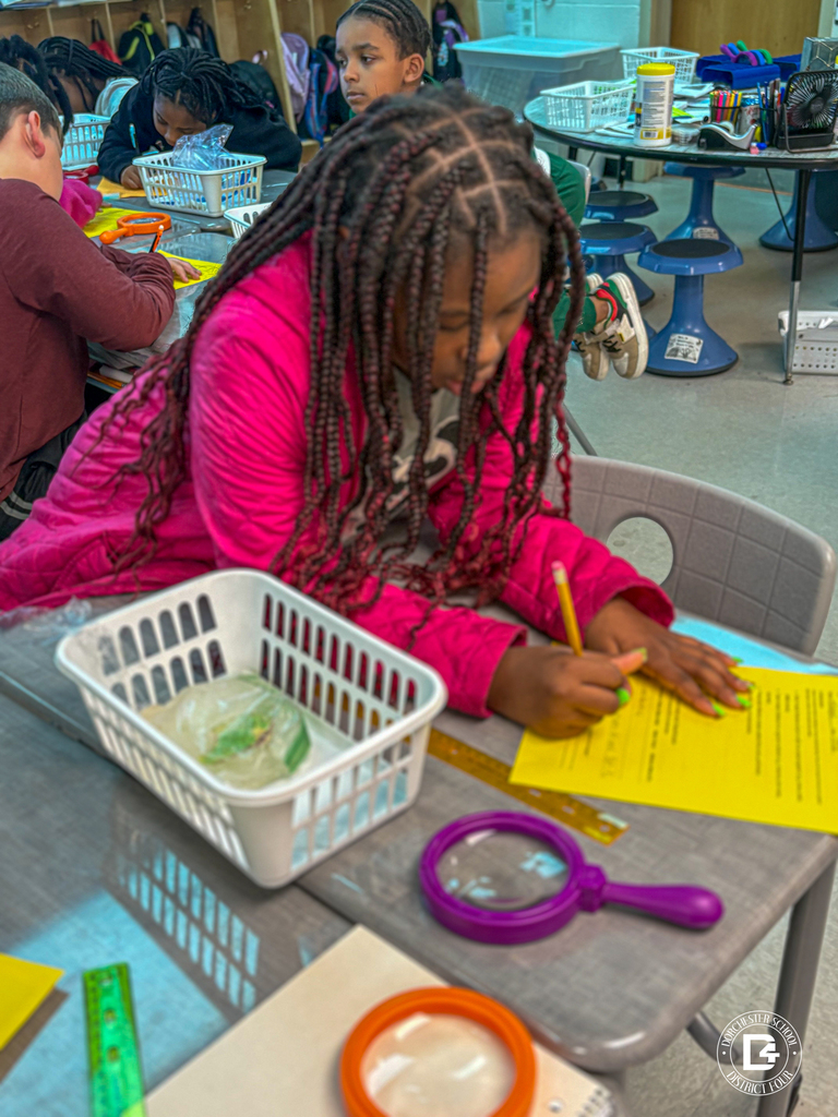 A third grade student in a pink jacket writes on a yellow worksheet while examining a classroom science sample. Magnifying glasses, rulers, and a basket with a frozen specimen are on the table. Other students work in the background.