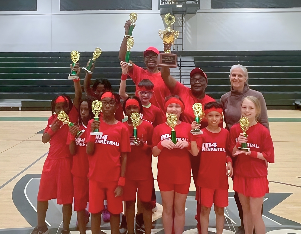 A group of elementary students in red “DD4 Basketball” uniforms stand on a gym floor holding gold trophies, smiling after winning their championship. Two adult coaches stand behind them, also holding a large trophy, while teammates raise smaller trophies in celebration. The team is gathered in front of bleachers, showing pride and excitement after their victory.
