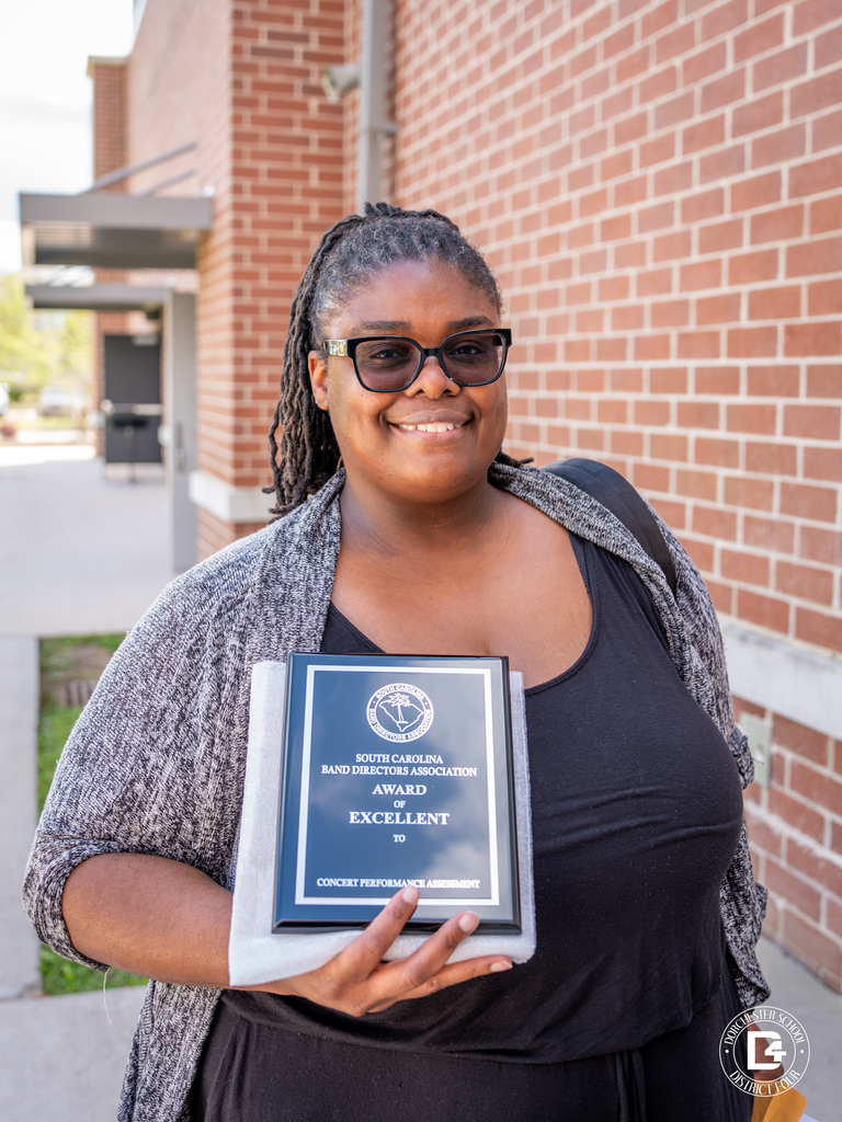  Ms. Yolanda Bennett stands outside near a brick wall, smiling while holding a plaque that reads “South Carolina Band Directors Association Award of Excellent” for Concert Performance Assessment. She is wearing glasses and a black outfit with a gray cardigan.