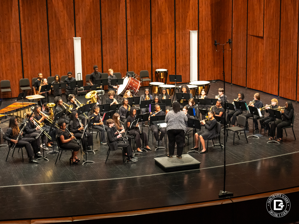 Woodland Middle School concert band performs on stage in a concert hall, dressed in black attire, seated in sections with instruments including flutes, clarinets, trumpets, trombones, and percussion. The band is led by director Ms. Yolanda Bennett, who stands on a podium conducting in front of the group, with wooden acoustic panels behind them and stage microphones set up.