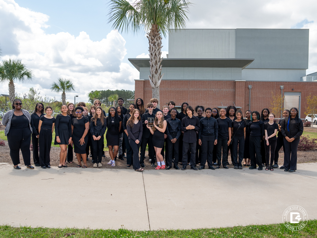 Woodland Middle School concert band members and director Ms. Yolanda Bennett pose together outside in front of the school building and palm trees. The group is dressed in black performance attire, with one student holding the award plaque at the center.