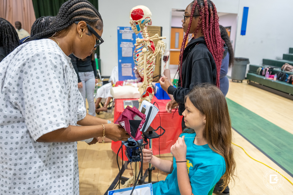 A WHS Health Science student, dressed in a hospital gown, demonstrates medical equipment to a visiting 8th grade student at the Health Science club table, with an anatomy skeleton displayed in the background.
