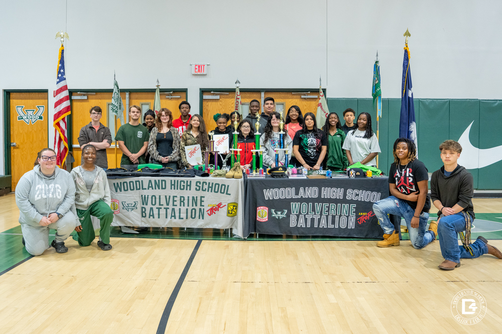 Members of the Woodland High School Army JROTC Wolverine Battalion pose for a group photo behind their display table, surrounded by trophies, flags, and recruitment materials.