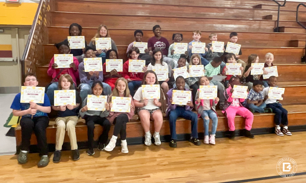 A large group of elementary school students sit on gymnasium bleachers at a Dorchester School District Four school, each proudly holding a "Student of the Month" certificate recognizing their character. The students, ranging in age from approximately 5–12, are arranged in several rows and smile at the camera. A teacher is visible among the group in the back rows. The gym floor and wooden bleachers are visible in the background. A Dorchester School District Four logo appears in the bottom right corner.