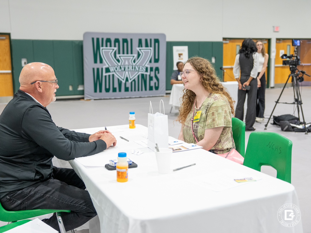 Student smiles during a one-on-one mock interview with a business professional, with a Woodland Wolverines backdrop visible behind them.
