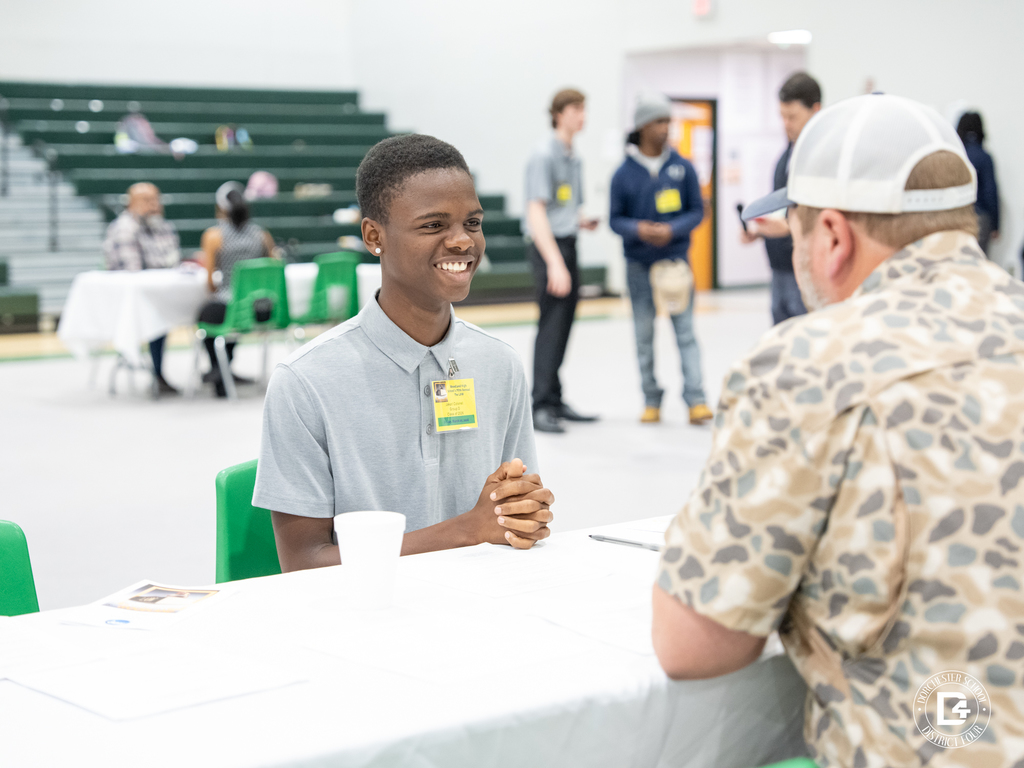 Student smiles while participating in a mock interview across from a volunteer in the gym, with other students and interview stations visible in the background.