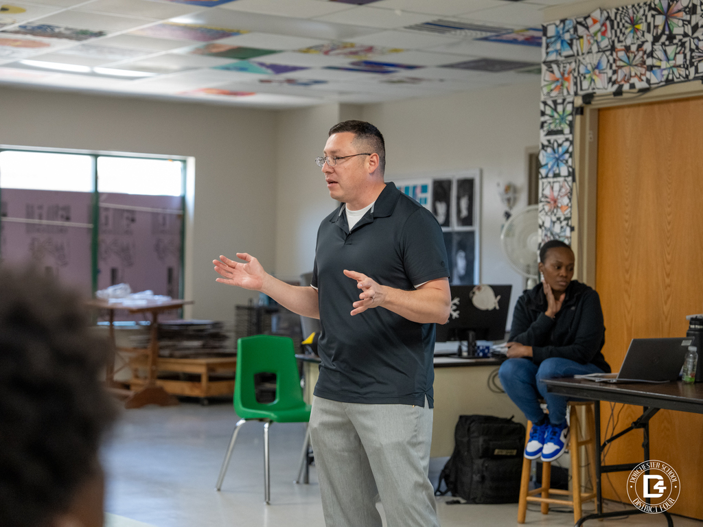 Presenter speaks to a group of students in a classroom setting, gesturing while another adult sits nearby with a laptop.
