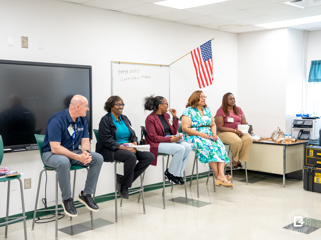 Panel of community members sits at the front of a classroom speaking with students during a session, with an American flag and classroom materials visible.