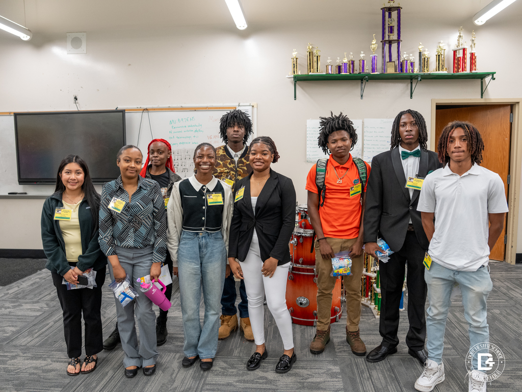 Group of student facilitators stand together in a classroom with trophies displayed behind them, dressed professionally and ready to support the event.