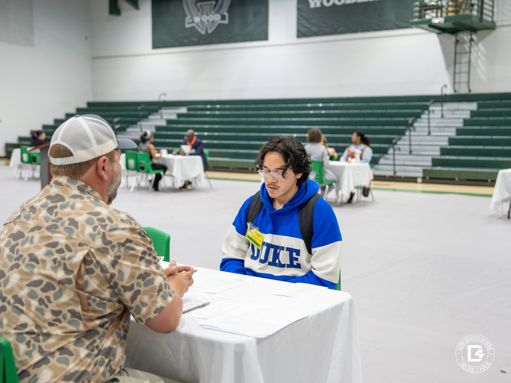 Student participates in a mock interview with a community volunteer inside Woodland High School’s gym, seated at a table with paperwork while other interviews take place in the background.