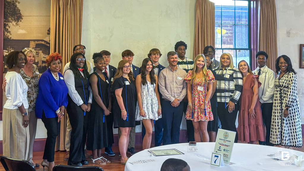 A large group of approximately 20 students and adult chaperones pose together smiling inside a banquet hall decorated with gold drapes and an American flag. Several students are wearing name tags and dressed in formal attire. A round table in the foreground displays a "Graduation Luncheon" centerpiece along with sponsor logos including HCA.