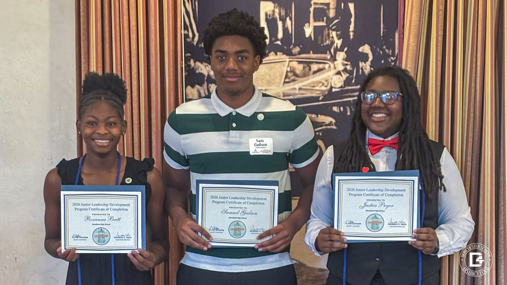 Three students stand side by side smiling and proudly holding their 2026 Junior Leadership Development Program Certificates of Completion from Morehead High School. The student on the left wears a black dress with a blue tassel, the student in the center wears a green and white striped polo with black pants and a name tag reading "Sam Gibson," and the student on the right wears a black suit with a red bow tie and dreadlocks.