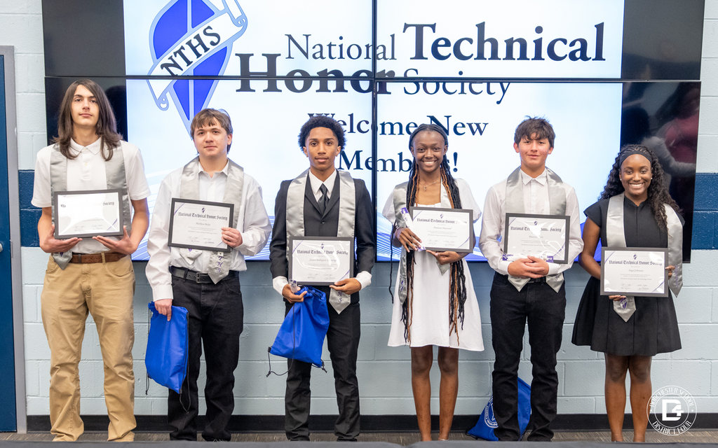 Six Dorchester School District Four students stand in a row in front of a large screen displaying "National Technical Honor Society – Welcome New Members!" Each student holds a framed National Technical Honor Society induction certificate and wears a silver NTHS stole. Some students also hold blue gift bags. The group is smiling and dressed in formal attire.