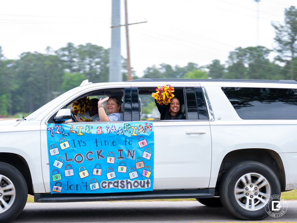 Staff ride in a white SUV decorated with a handmade testing-themed sign that reads, “It’s time to lock in, don’t crashout,” while waving pom-poms from the windows.