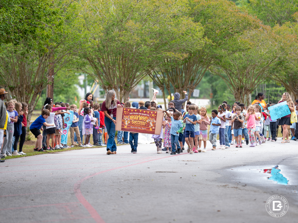 Students and staff line a school driveway during a parade, with children holding signs and walking together under trees.